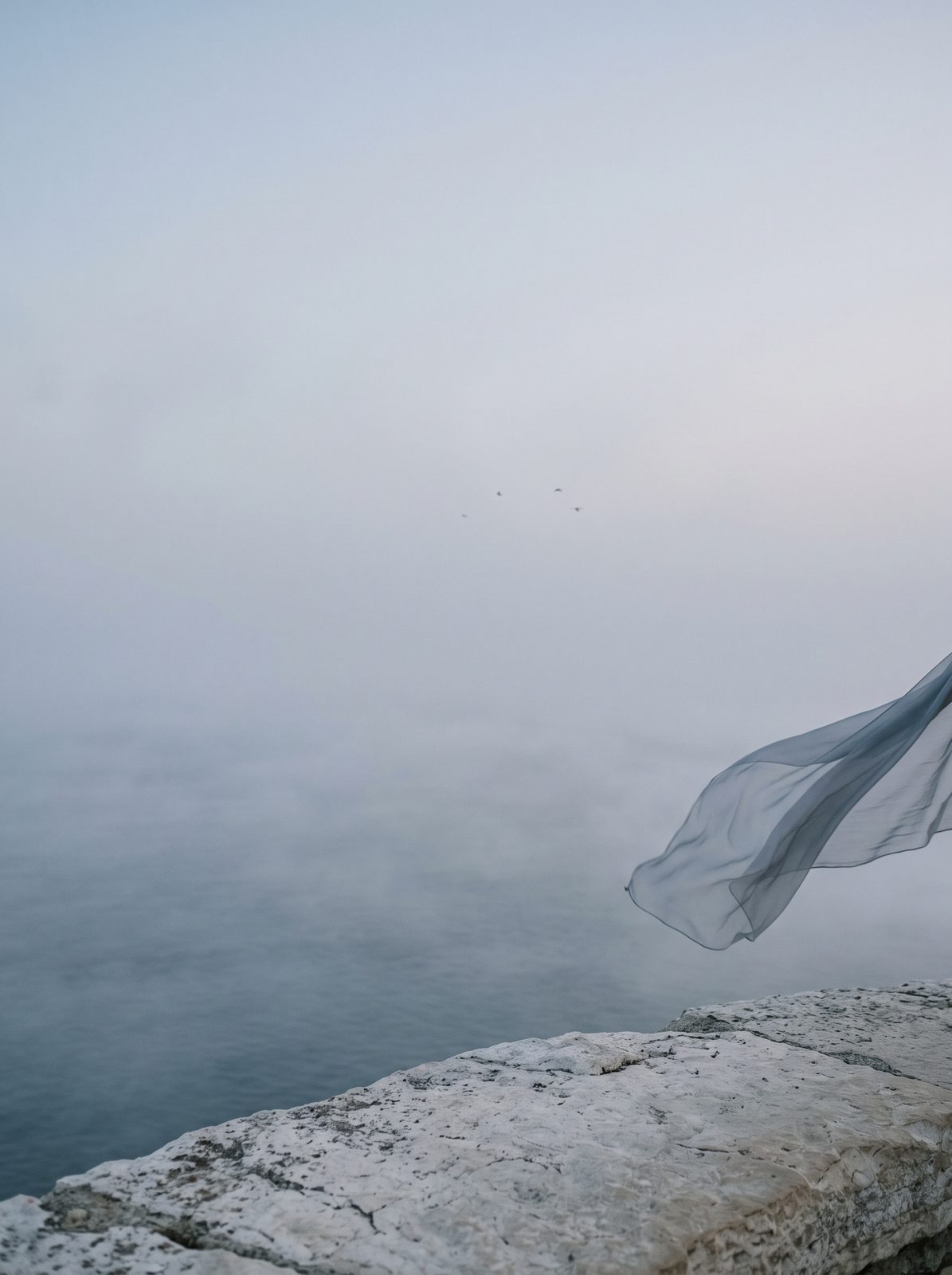 A misted sea at blue hour, a silk scarf lifting in the wind